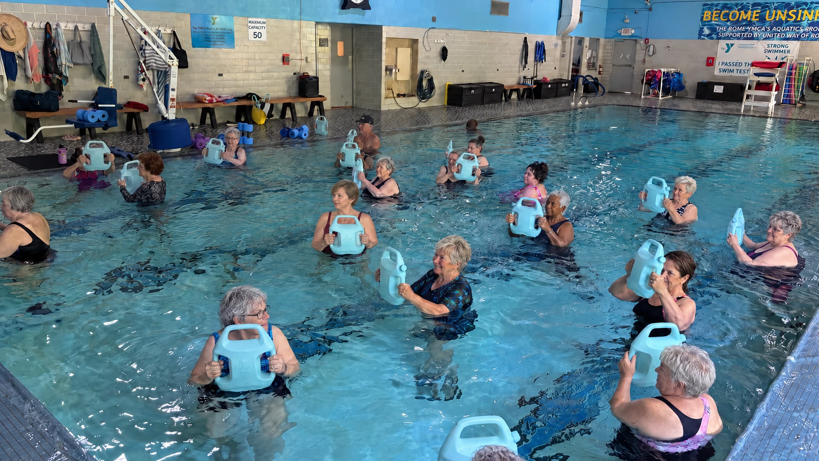 Older adults in an indoor pool aqua fitness class, each holding an AguaForce water weight at chest height